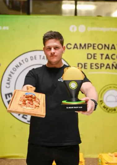 Hombre sosteniendo un trofeo y una placa en el podio de una competencia de tacos españoles, con un fondo amarillo para el evento.
