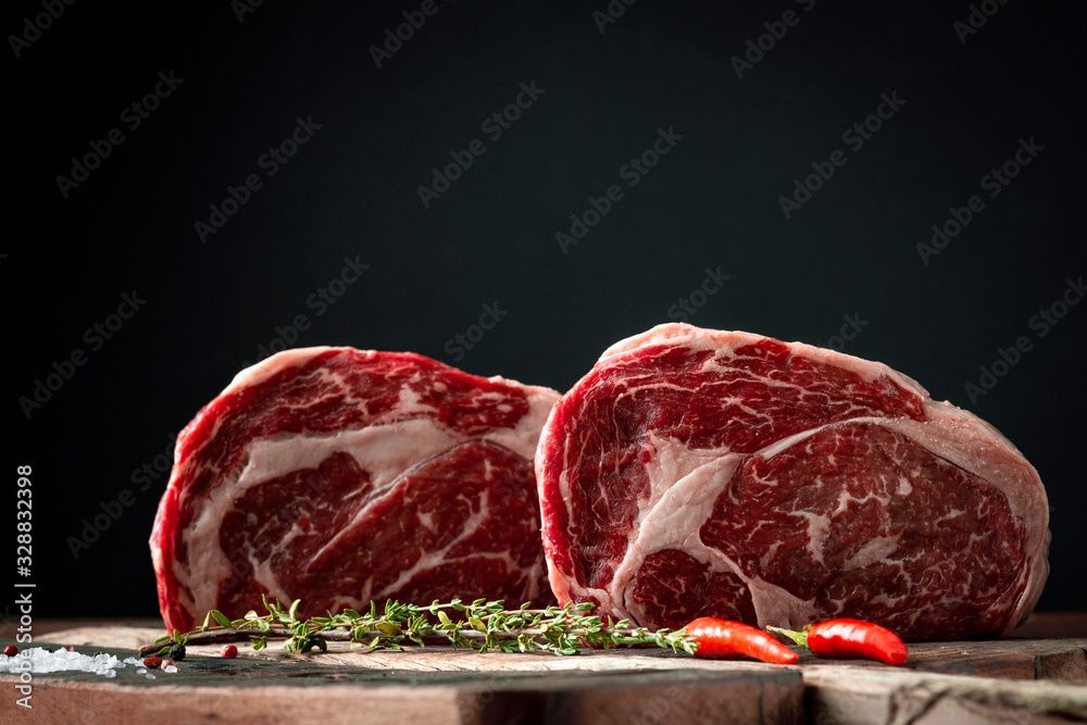 Zwei rohe Rindersteaks auf einem Holzbrett mit Kräutern und Kirschtomaten vor dunklem Hintergrund