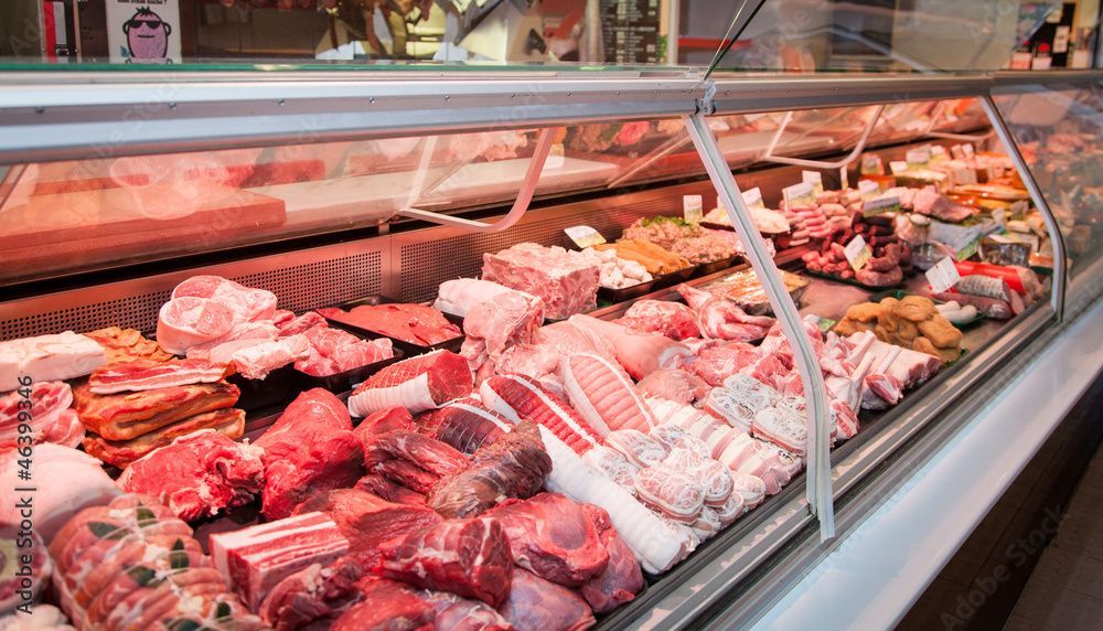 Fleischtheke mit verschiedenen rohen Fleischsorten und Wurstwaren, die unter Glas in einem Markt ausgestellt sind.