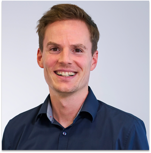 Lächelnder Mann mit kurzen braunen Haaren, trägt ein blaues Button-Down-Hemd und blickt in die Kamera. Grau-weißer Hintergrund.