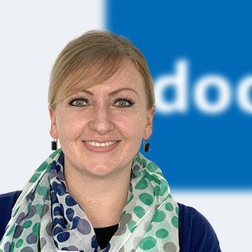 Frau mit blondem Haar und Schal lächelt vor einem blau-weißen Hintergrund.