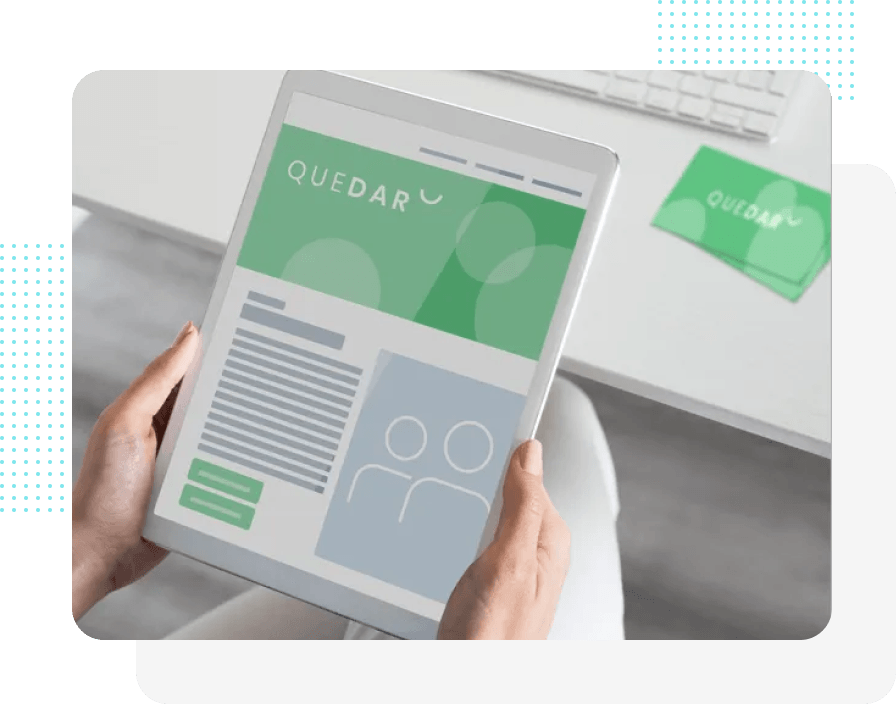 Person hält ein Tablet mit einer Website und dem Logo „QUEDAR“ auf einem weißen Schreibtisch mit einer Visitenkarte.