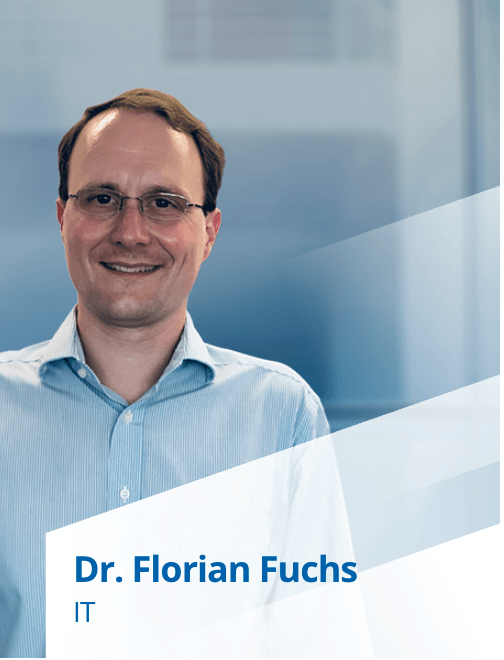 Dr. Florian Fuchs, ein IT-Experte, lächelt in einem hellblauen Hemd. Im Hintergrund ist ein unscharfer Büroraum zu sehen.