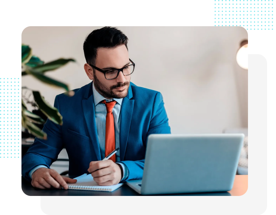 Mann in blauem Anzug mit roter Krawatte, Brille, schaut auf einen Laptop und schreibt in ein Notizbuch an einem Schreibtisch.