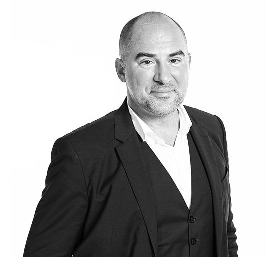 Lächelnder Mann mit Brille, graues Hemd, vor blau-weißem Hintergrund.