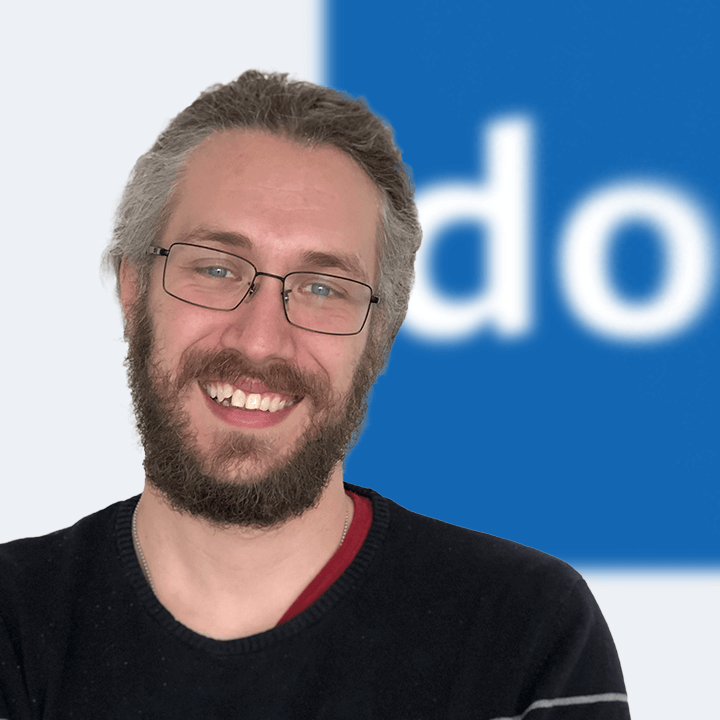 Mann mit Brille und Bart lächelt, blau-weißer unscharfer Hintergrund.