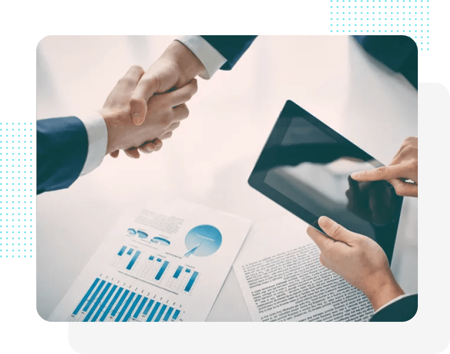 Zwei Menschen in Anzügen schütteln sich über einem Tisch mit Diagrammen und einem Tablet die Hände.