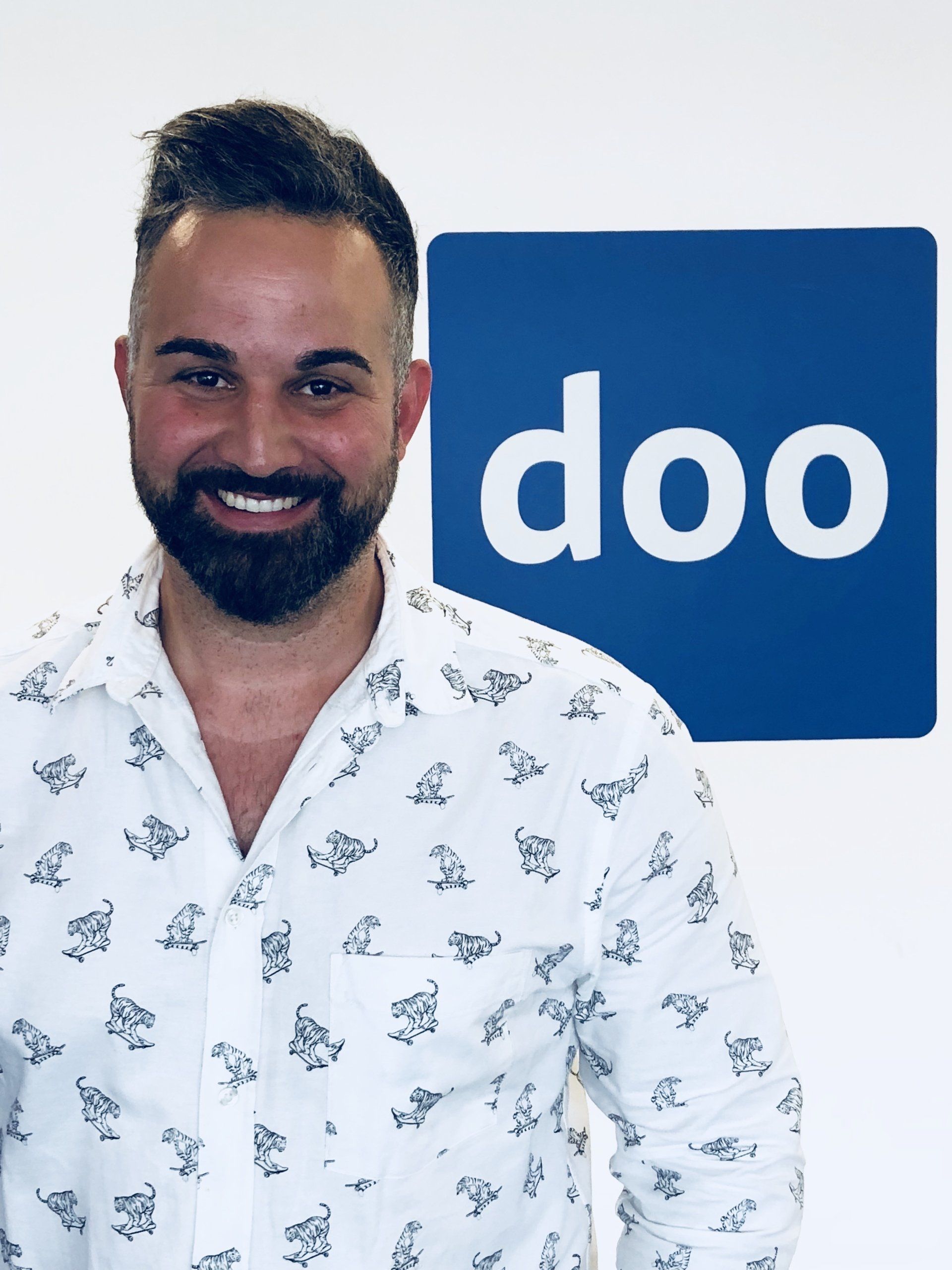 Lächelnder Mann mit Brille, graues Hemd, vor blau-weißem Hintergrund.