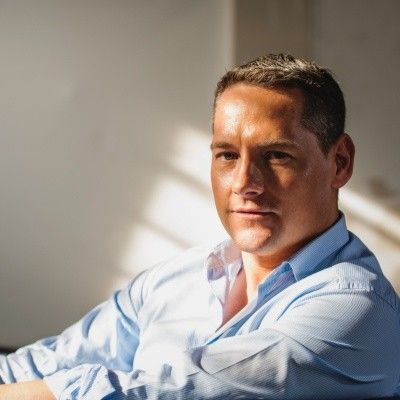 Lächelnder Mann mit Brille, graues Hemd, vor blau-weißem Hintergrund.