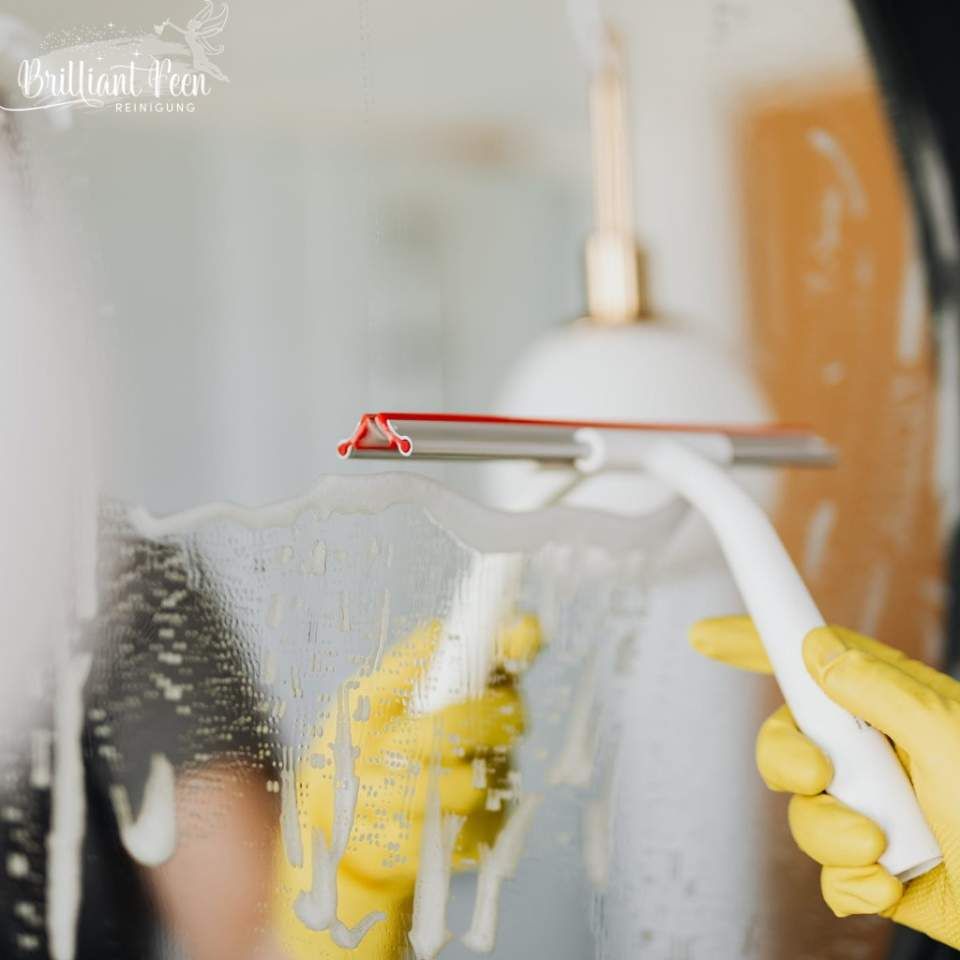 Eine Person mit gelben Handschuhen benutzt einen Abzieher, um einen seifigen Spiegel zu reinigen.