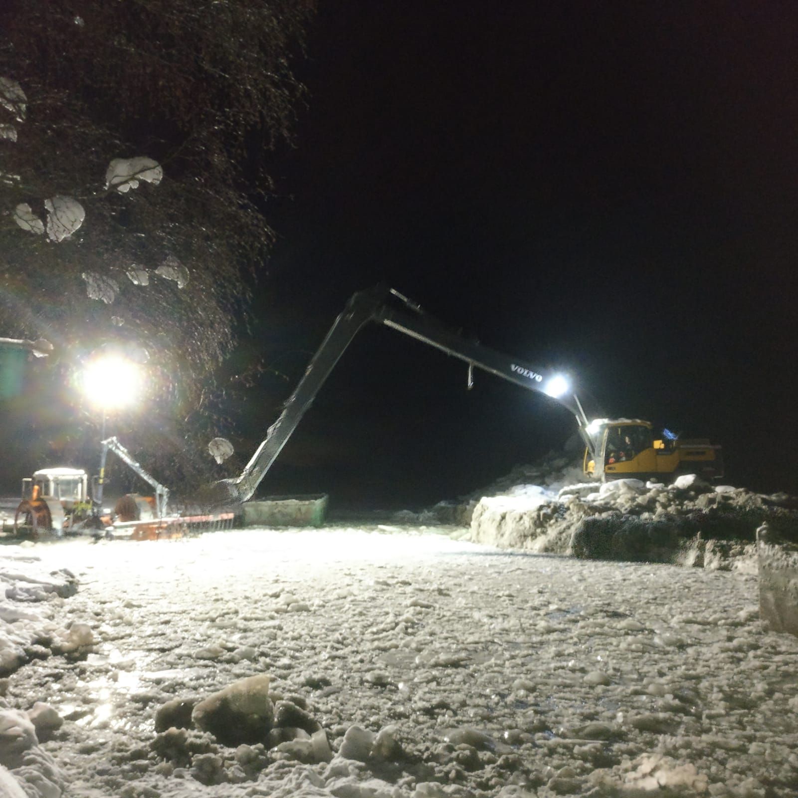 Yöllistä lumenaurausta pitkällä kaivinkoneella ja kuorma-autoilla kirkkaiden työvalojen alla.