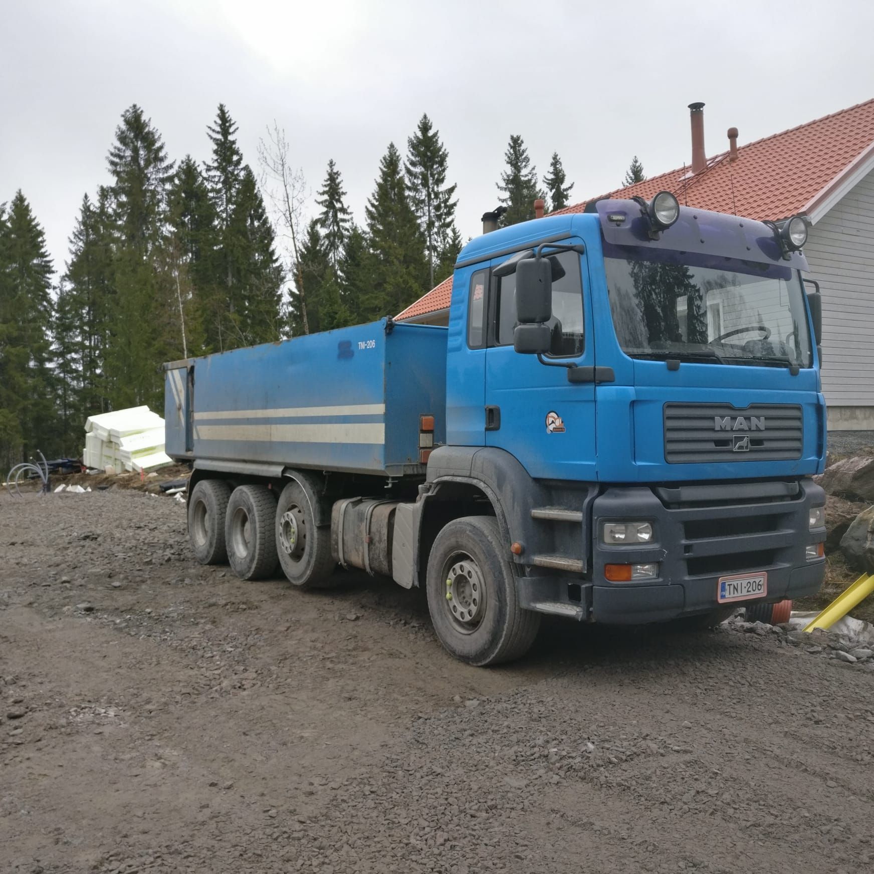 Sininen MAN-kippiauto pysäköitynä mutaiselle tontille punakattoisen rakennuksen ja mäntyjen viereen