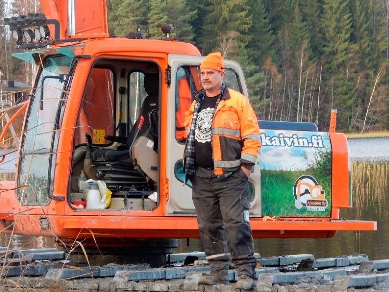 Oranssi kaivinkone lumisella työmaalla, työntekijä seisoo sen vieressä ja metsä taustalla