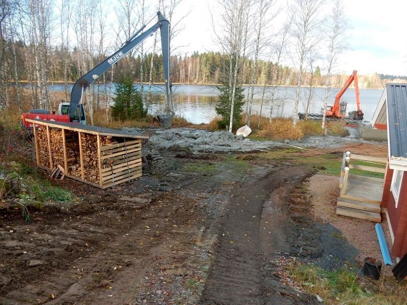 Rakennustyömaa, jossa kaivinkoneita järven rannalla, hiekkatie ja pinottuja materiaaleja puiden lähellä