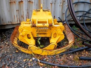 Keltainen hydraulinen purkulaite soralla mustien letkujen ja ruostuneen metallisäiliön vieressä.