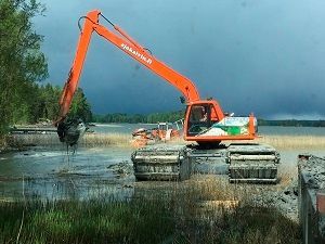 Oranssi kaivinkone ponttoneilla työskentelee tulvivalla suolla pilvisen taivaan alla