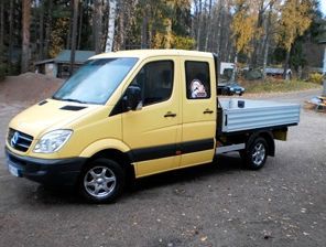 Keltainen lavetilla varustettu hyötykuorma-auto pysäköitynä hiekkatielle puiden viereen