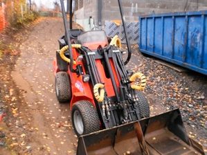 Punainen ruohonleikkuri etukuormaajalla ja taitetuilla lisälaitteilla lehtien peittämällä polulla sinisen roskalavan vieressä
