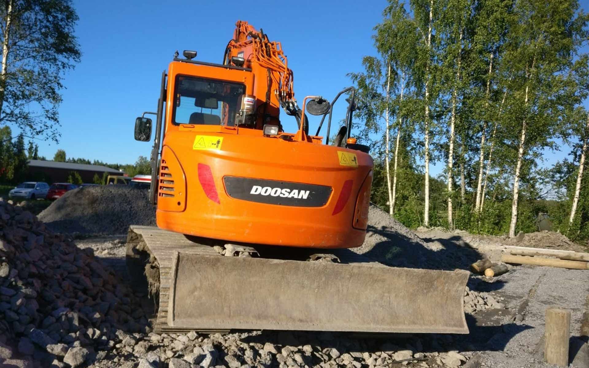 Oranssi Doosan-kaivinkone maakasalla rakennustyömaalla, taustalla puita ja sinistä taivasta
