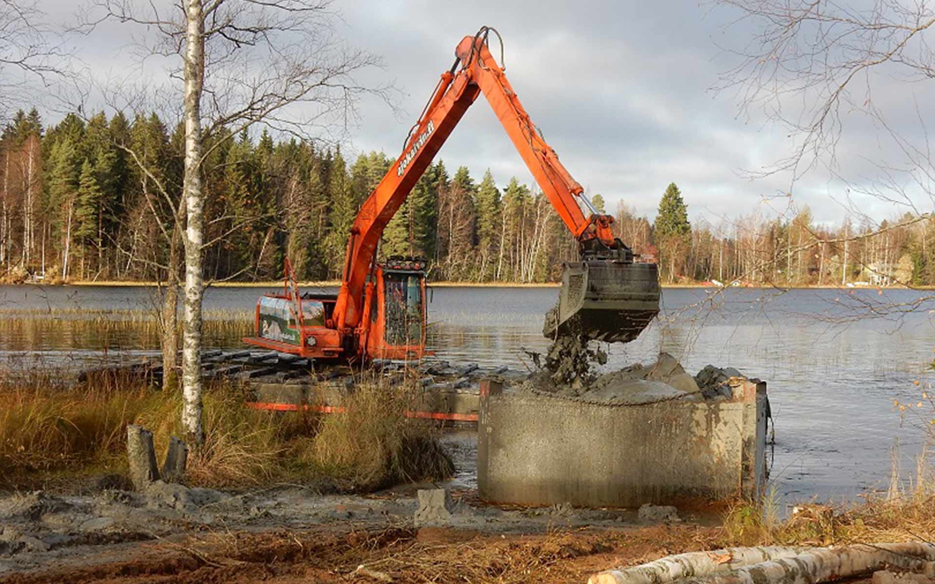 Oranssi kaivinkone kauhaa roskia betonista joessa, taustalla metsää ja rantaviivaa