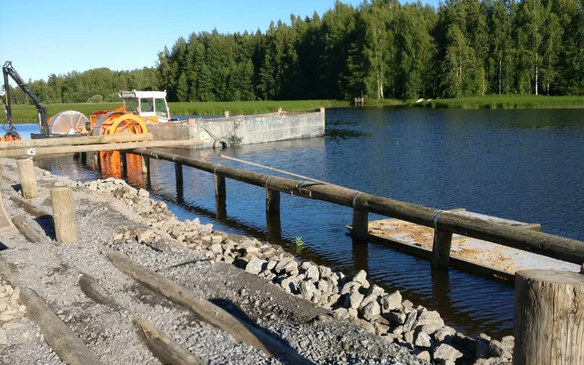 Rakennustyömaa järven rannalla, jossa on soraranta, puiset kaiteet ja metsäinen tausta