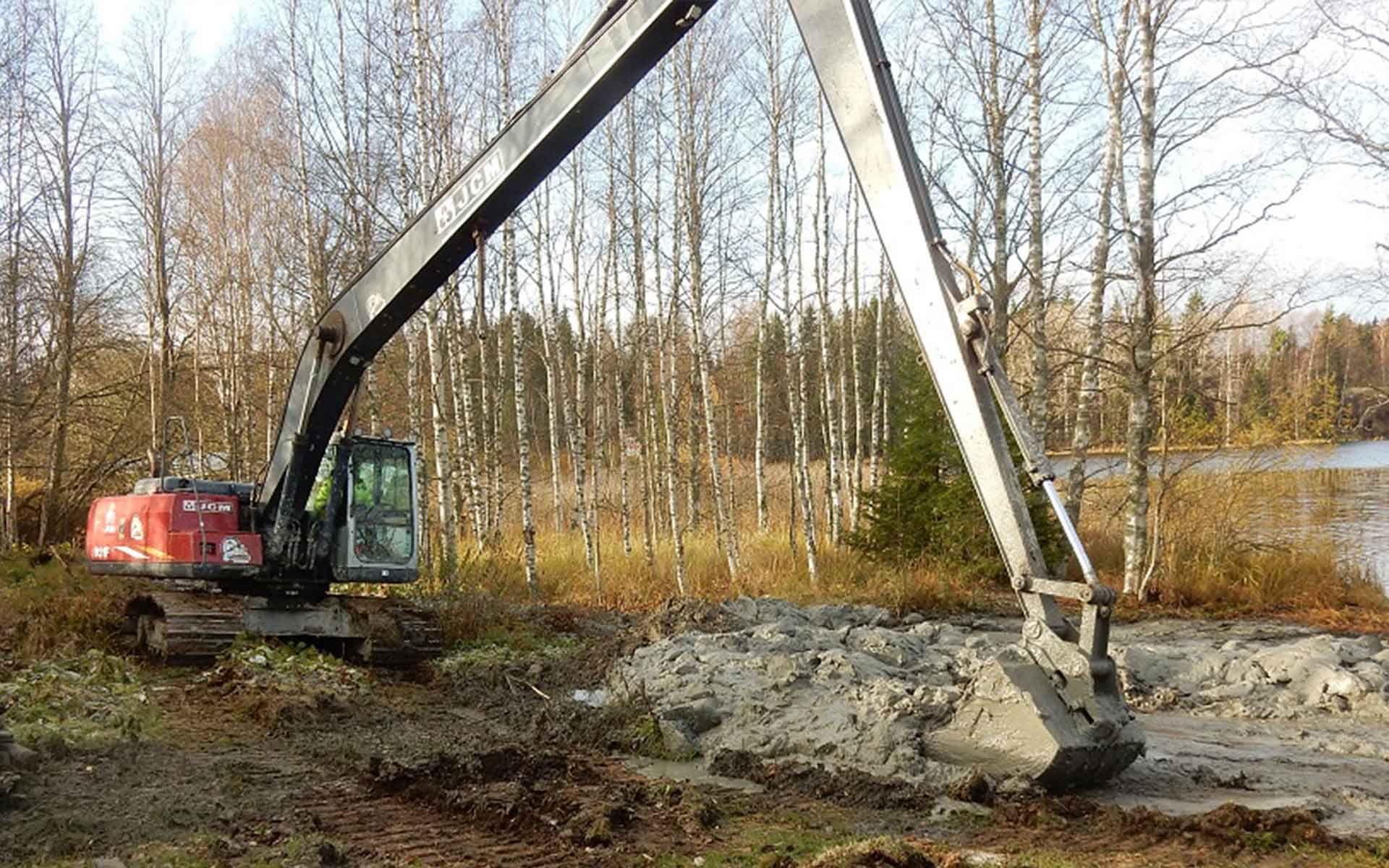 Pitkällä puomilla varustettu kaivinkone kaivaa mutaista maata metsäisen lammen lähellä