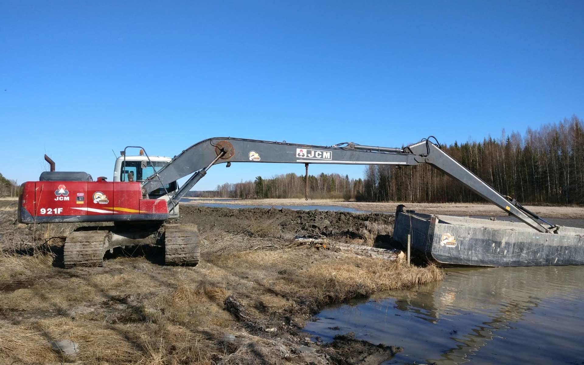 Punainen kaivinkone mutaisella rakennustyömaalla veden äärellä, käsivarsi ojennettuna betoniseinän yli sinisen taivaan alla