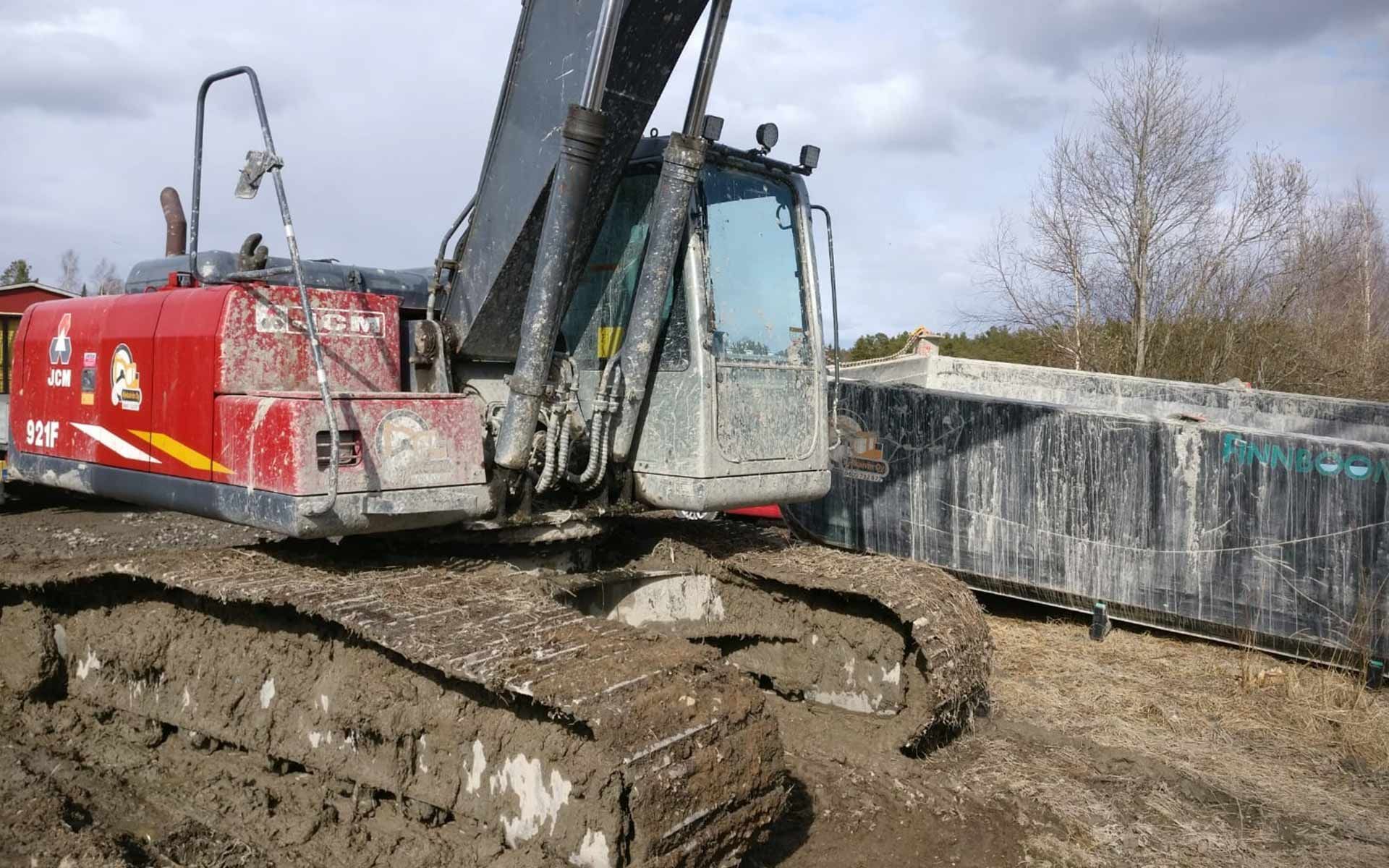 Kaivinkone kaivaa mutaista maata betoniseinän ja punaisen teollisuuskontin vieressä.