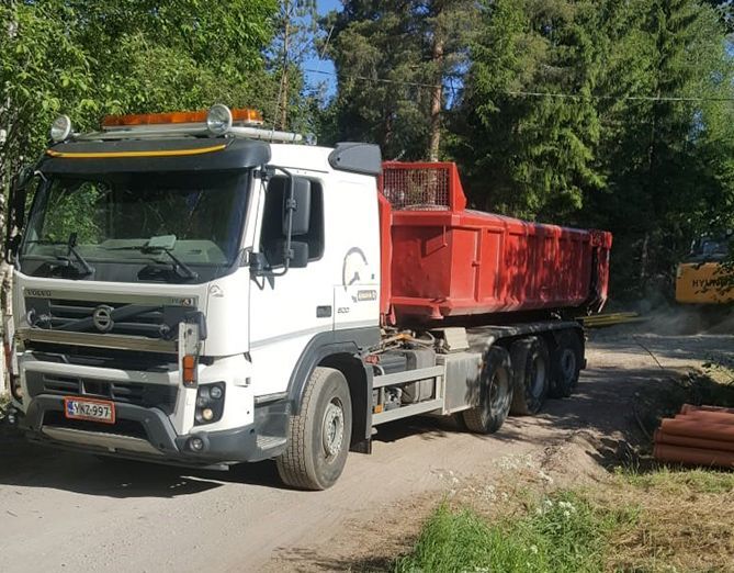 Valkoinen Volvo-kippiauto, jossa on punainen lava, pysäköitynä hiekkatielle puiden lähelle