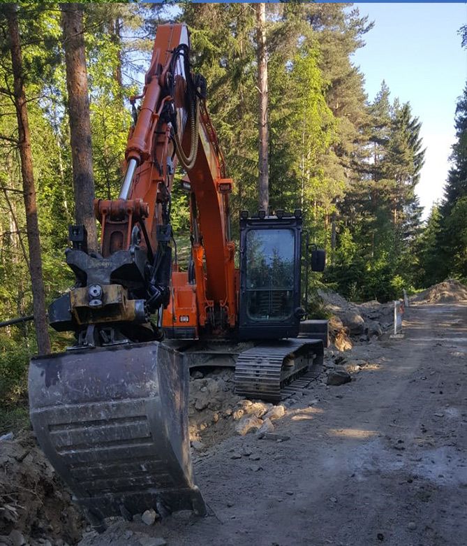 Oranssi kaivinkone hiekkatiellä metsäaukeamalla, korkeiden mäntyjen ympäröimänä.