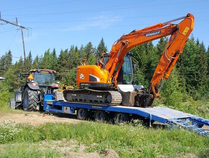 Oranssi kaivinkone sinisellä lavetilla tienvarrella nurmialueella traktorin ollessa lähellä.
