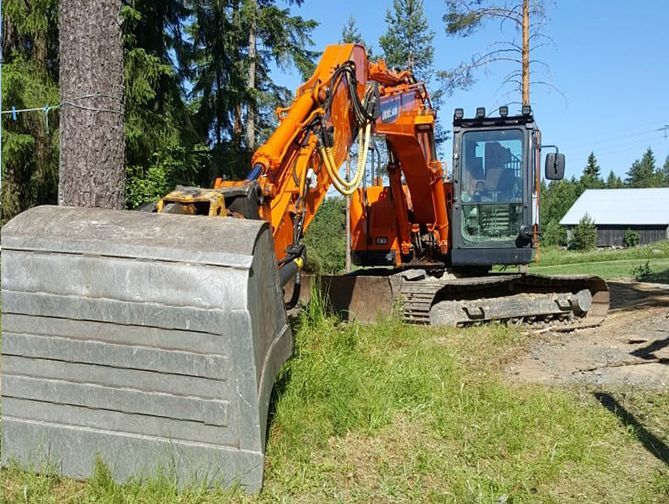 Oranssi kaivinkone nostaa suurta betoniseinäpaneelia nurmikentällä talon vieressä.