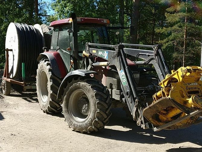Punainen traktori etukuormaajalla vetää letkukelakärryä hiekkatiellä metsäalueella