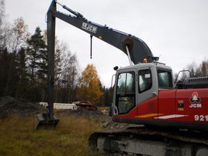 Punainen JCM-kaivinkone nostaa kauhaa nurmikentällä puiden vieressä ja pilvisellä taivaalla