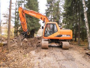 Oranssi kaivinkone raivaa likaa metsätiellä korkeiden puiden vieressä