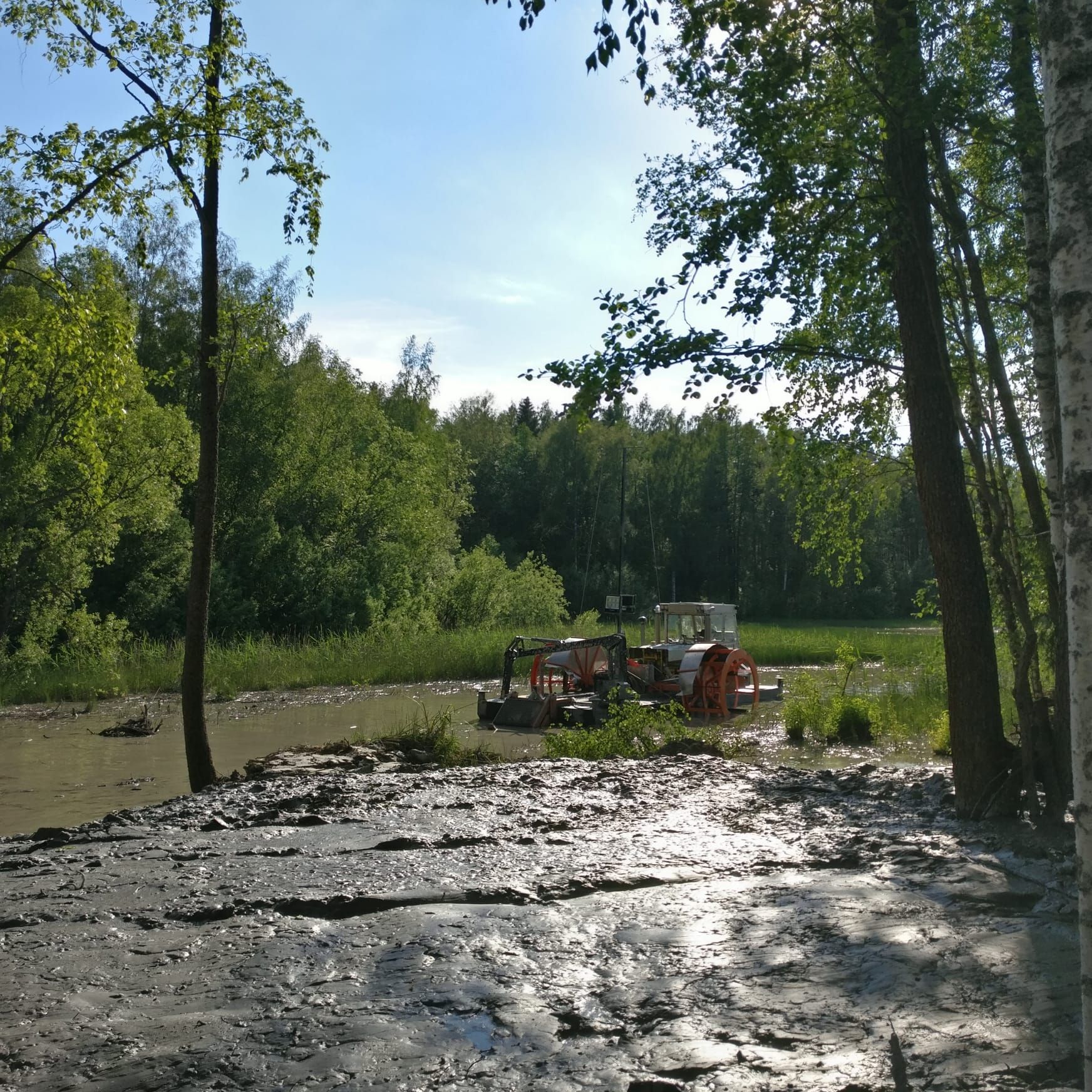 Mutainen joenranta, pieni oranssi traktori puiden lähellä ja aurinkoinen metsäaukeama