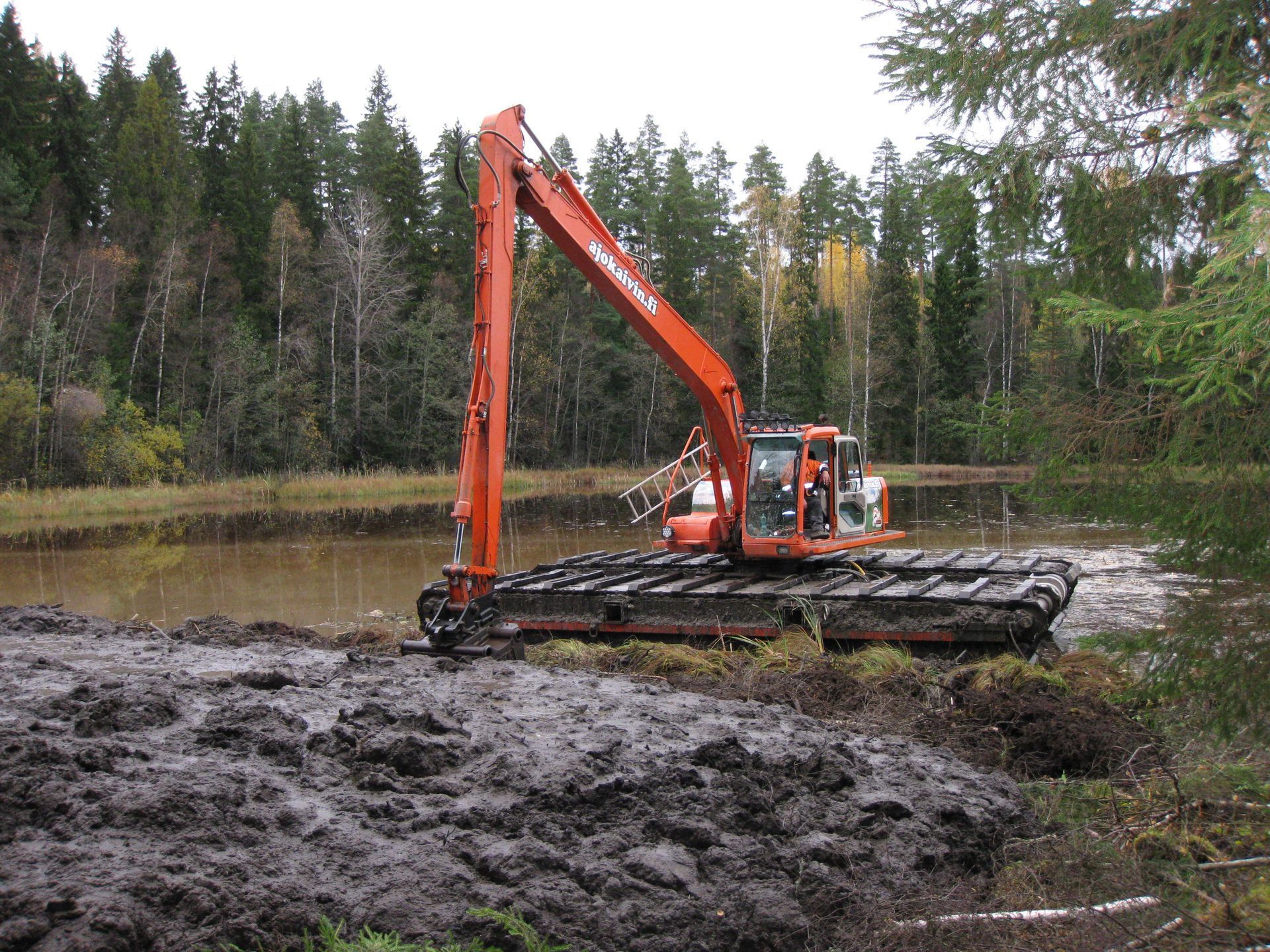 Oranssi kaivinkone kiskoilla työskentelee mutaisella kosteikkoalueella metsälammen vieressä