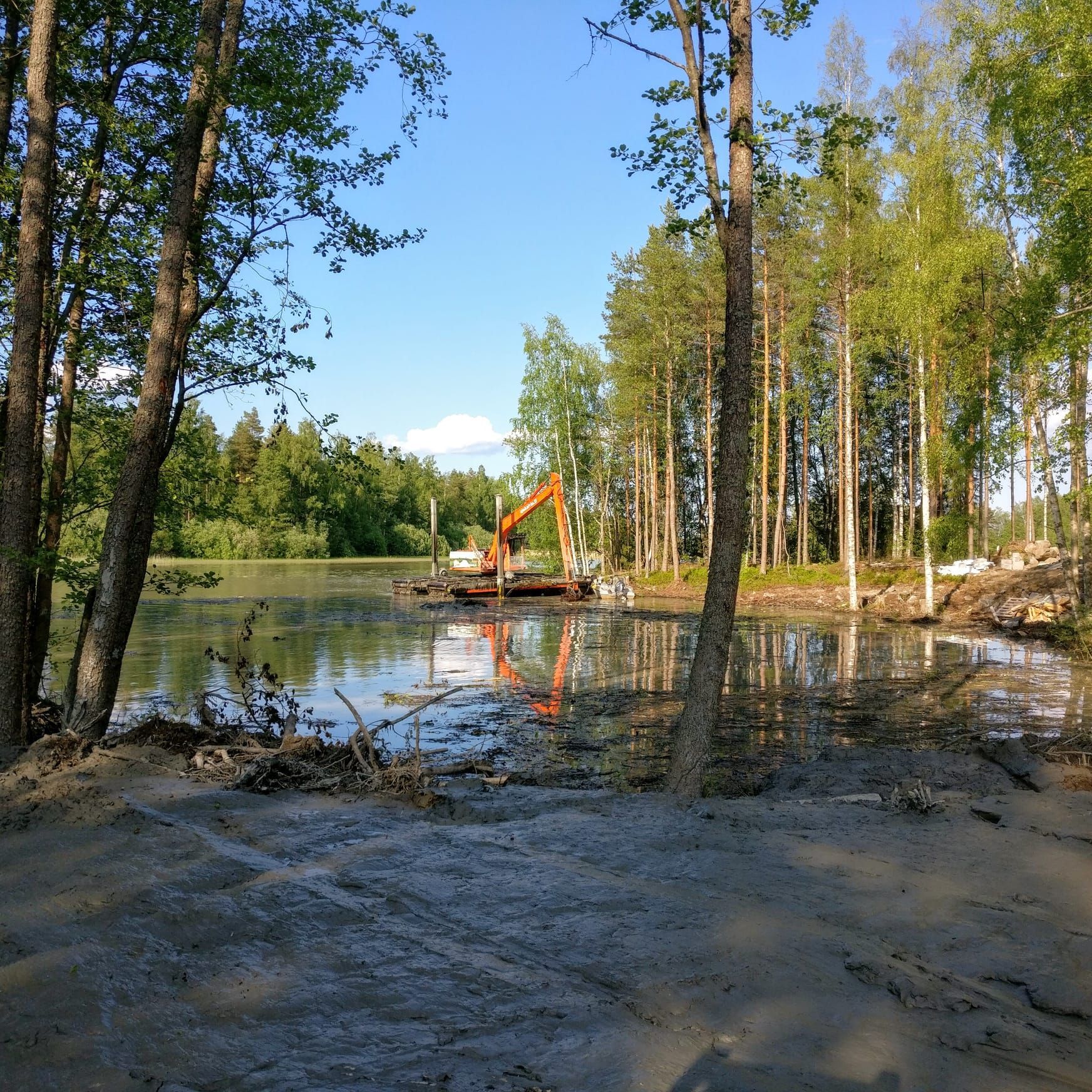 Oranssi kaivinkone lammen rannalla metsäalueella, puiden heijastuessa vedessä.
