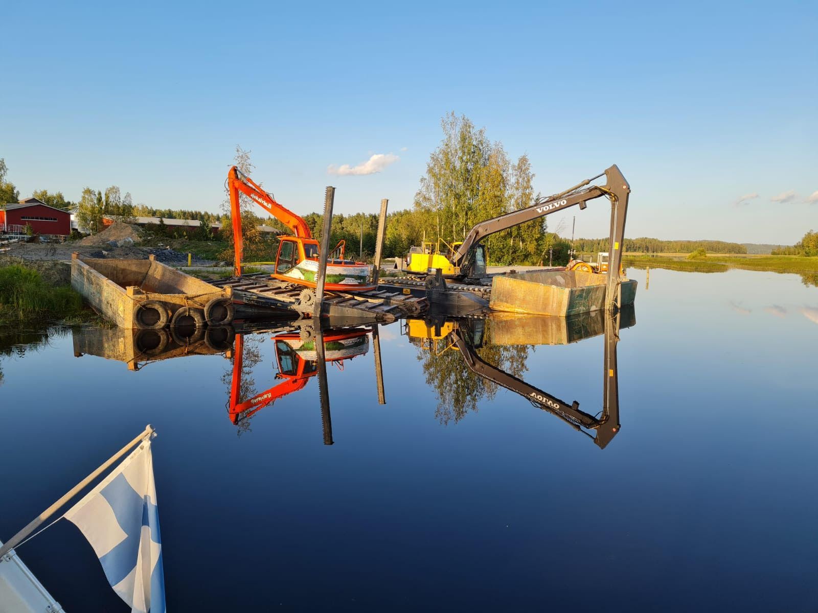Kaivinkoneita ja proomuja tyynellä joella, kirkkaat heijastukset ja sinivalkoinen lippu etualalla