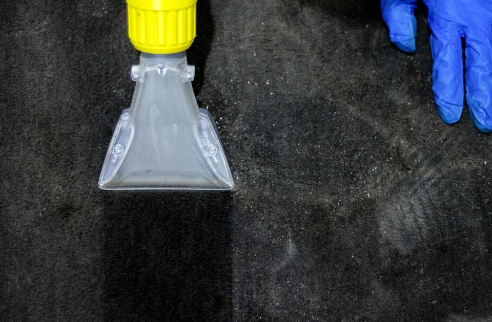 Eine Person mit blauen Handschuhen reinigt einen schwarzen Teppich mit einem Staubsauger.