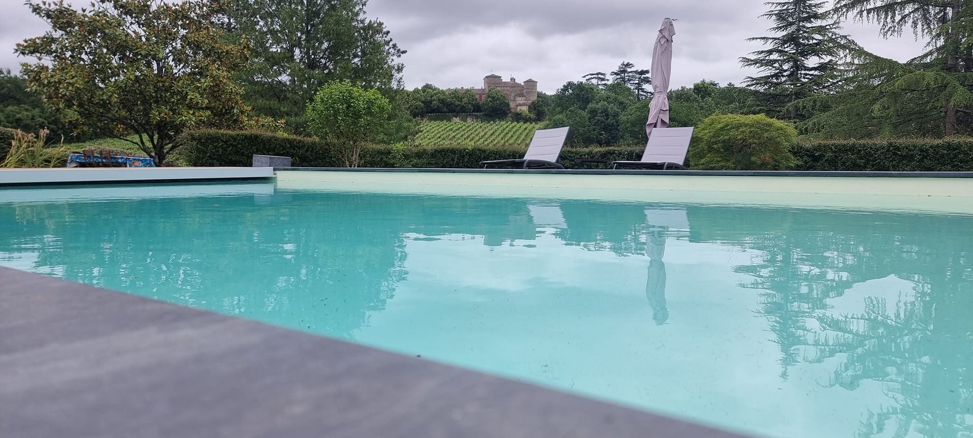 Le bord d'une piscine, un château est visible dans le fond derrière les haies végétales.
