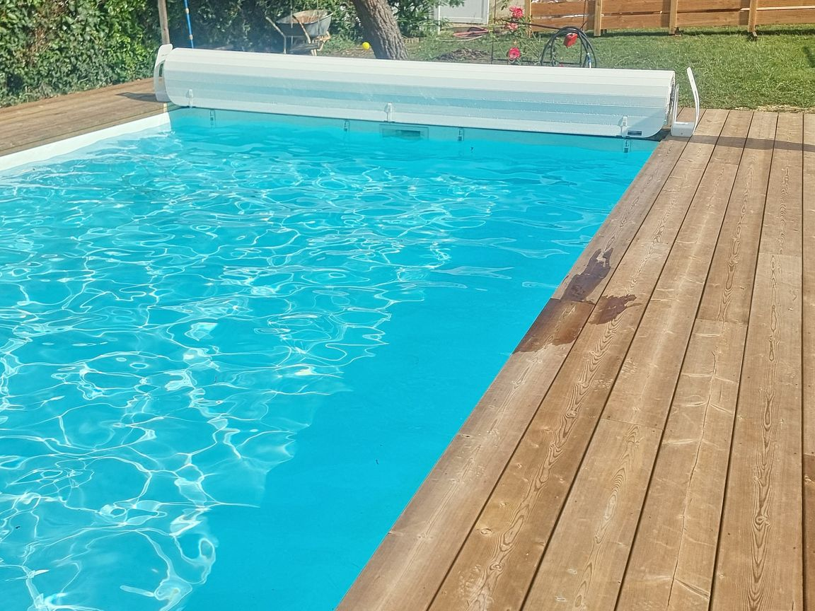 Piscine avec couverture blanche, entourée d'une terrasse en bois et d'une pelouse verte.
