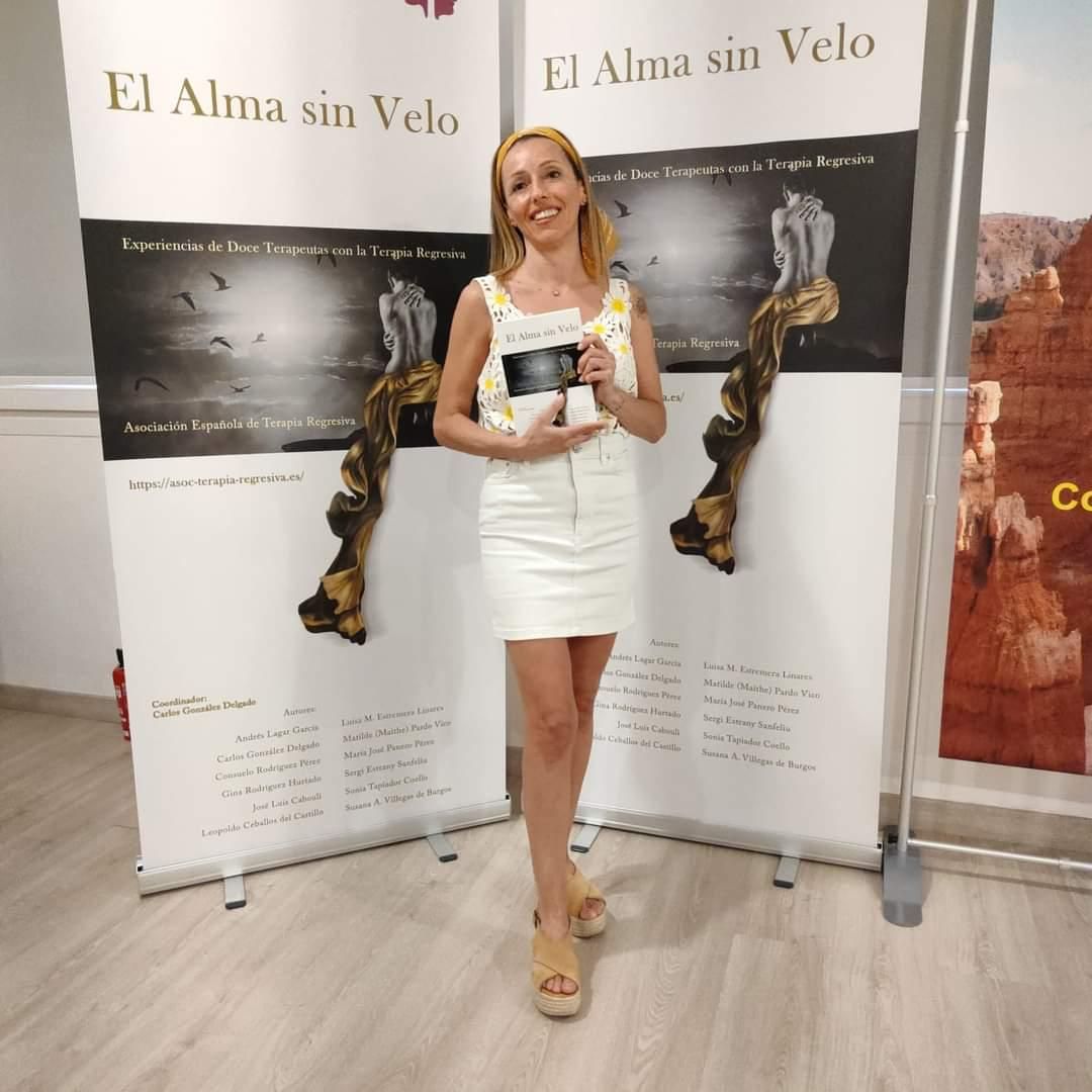 Mujer vestida de blanco sosteniendo un trofeo en un stand de un evento con pancartas de “El Alma sin Velo”.