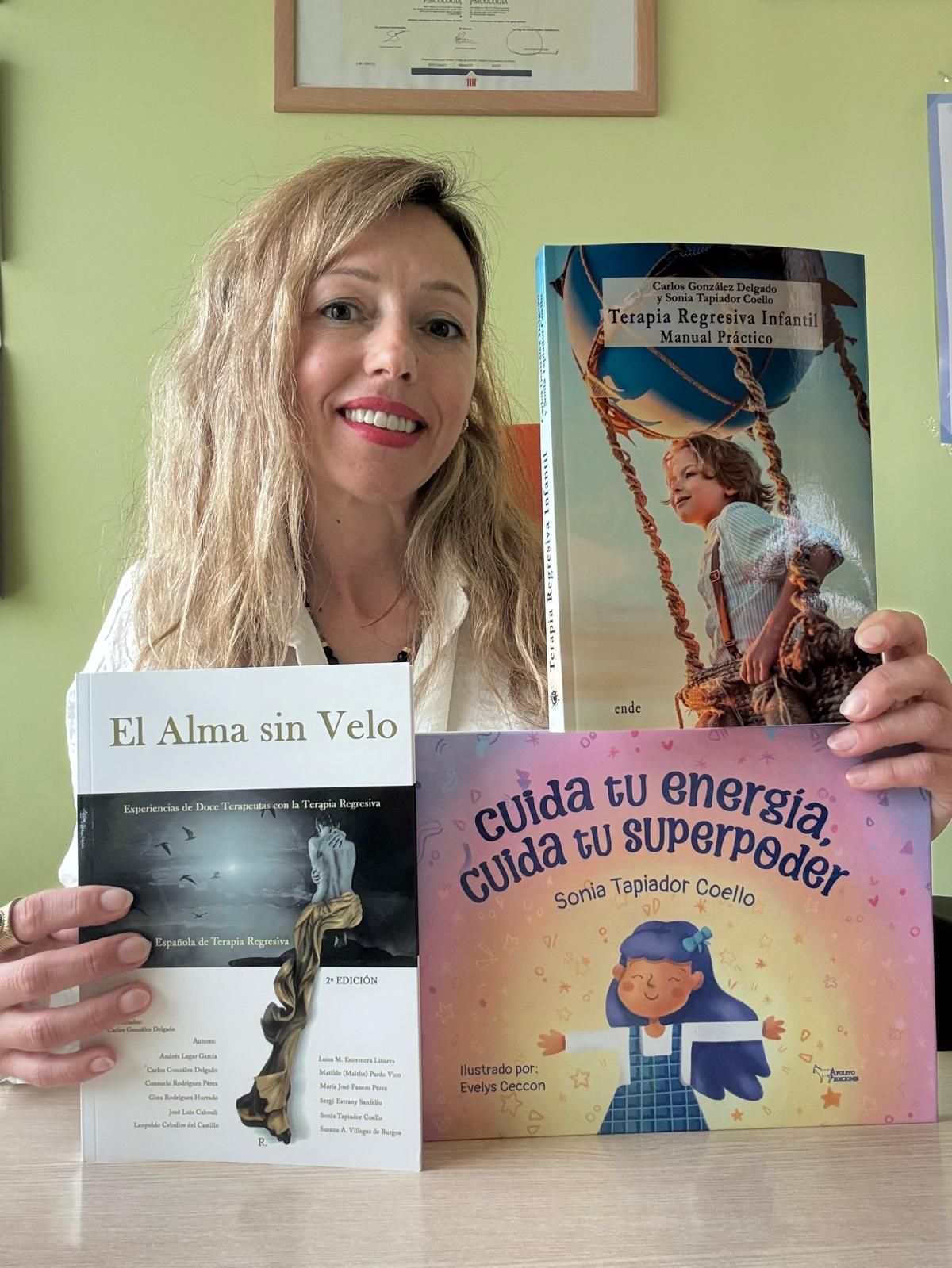 Mujer sosteniendo tres libros ilustrados, incluyendo uno titulado “Fi Alma sin Vuelo”, contra una pared verde claro.