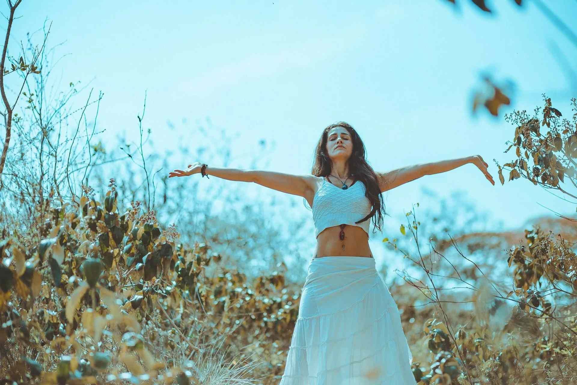 Una mujer con falda blanca y blusa azul permanece de pie con los brazos extendidos en un campo soleado.