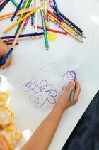 Manos coloreando una flor y pequeñas caras sobre papel blanco, con muchos lápices de colores esparcidos por encima.