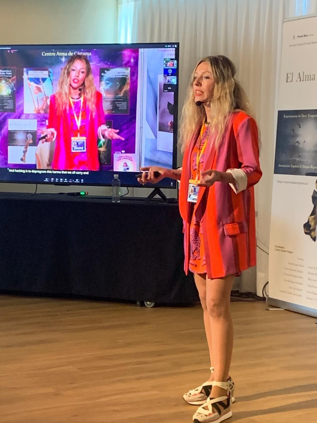 Mujer con un atuendo naranja brillante presentando junto a una gran pantalla que muestra su imagen en un evento.