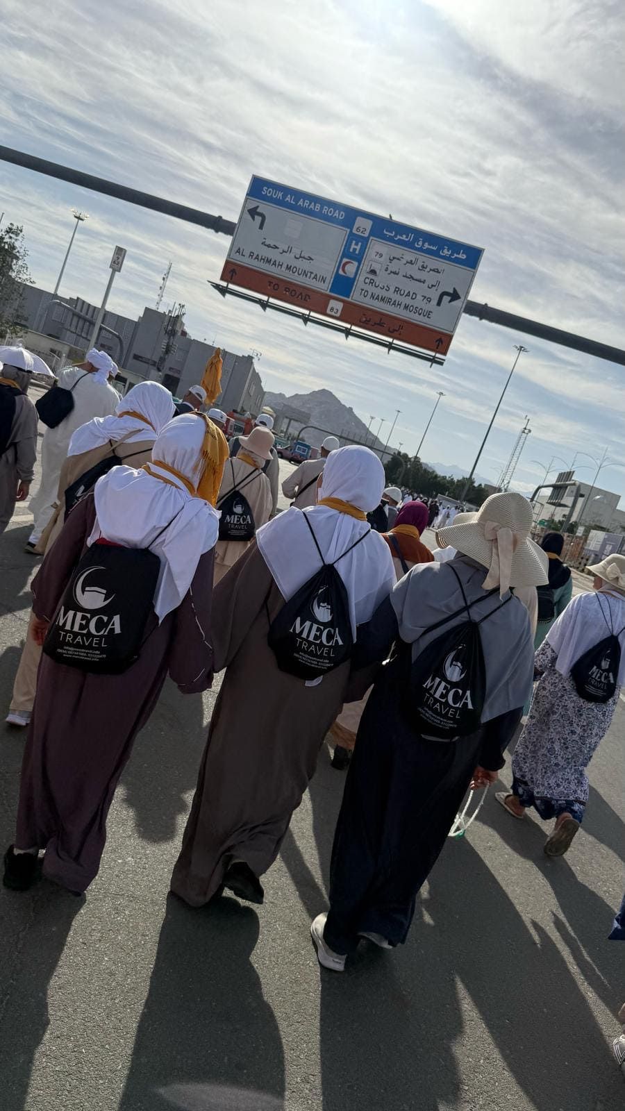 Peregrinos con túnicas blancas y marrones y mochilas caminan por una calle de La Meca, siguiendo señales direccionales.