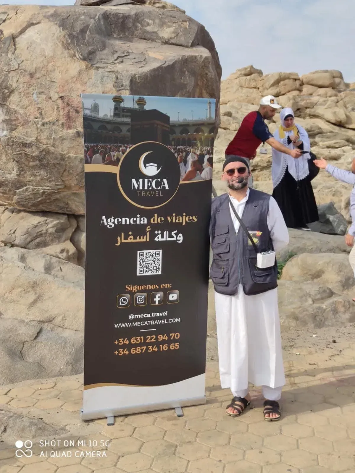 Hombre vestido de blanco de pie junto a un letrero de agencia de viajes con imágenes de La Meca, en un entorno al aire libre.