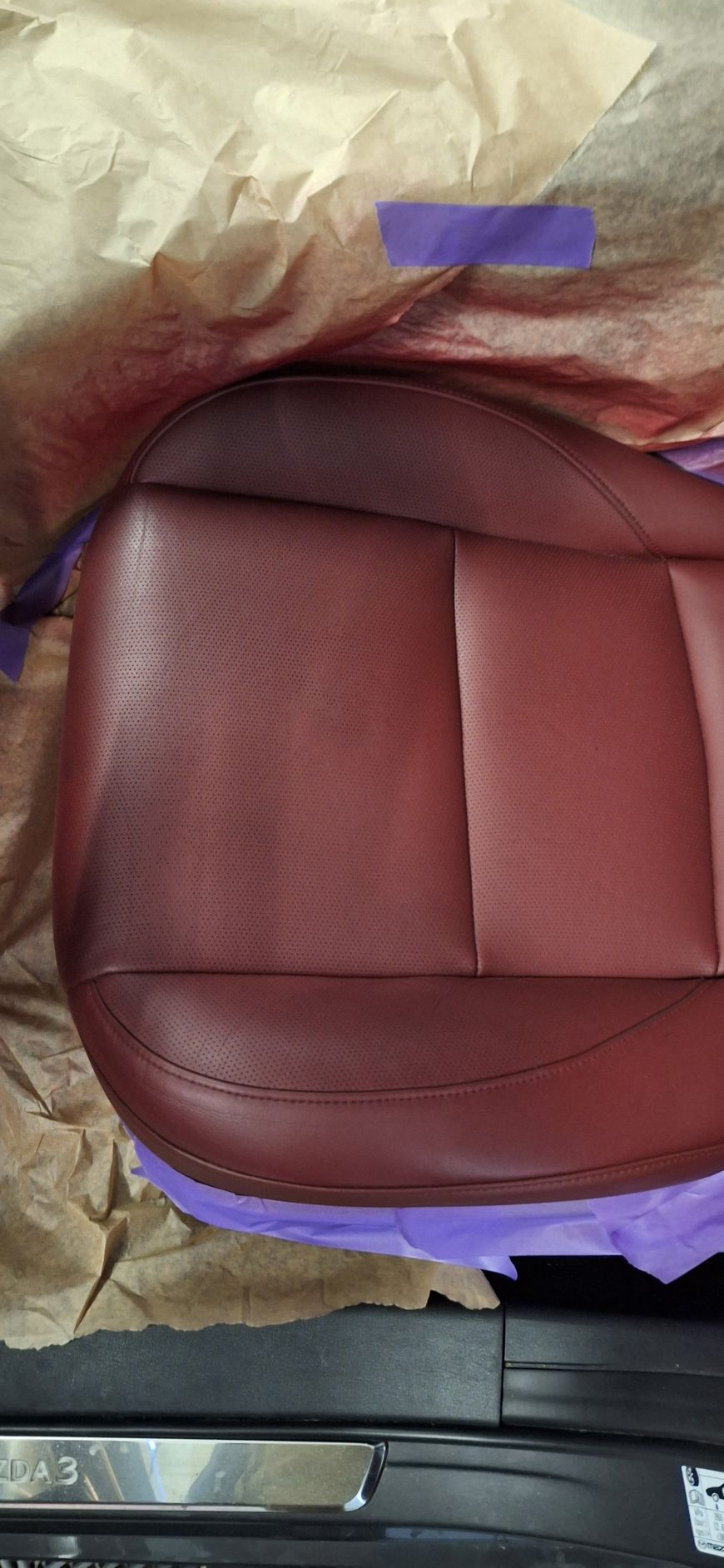 Gros plan sur un siège de voiture en cuir bordeaux avec des détails perforés, partiellement recouvert de ruban adhésif et de papier violet.